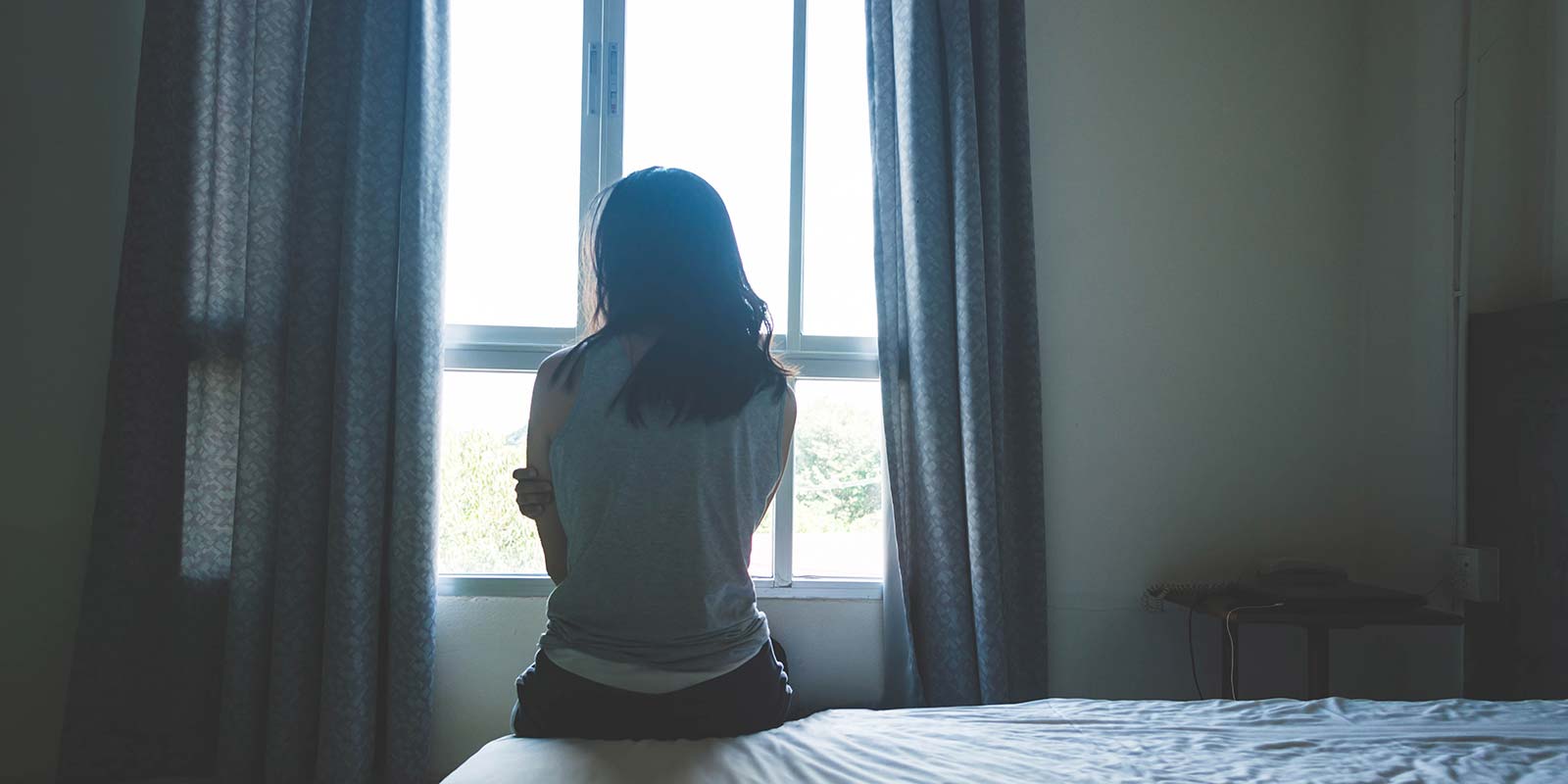 a woman in a dark woman next to a window, sitting on a bed, back turned